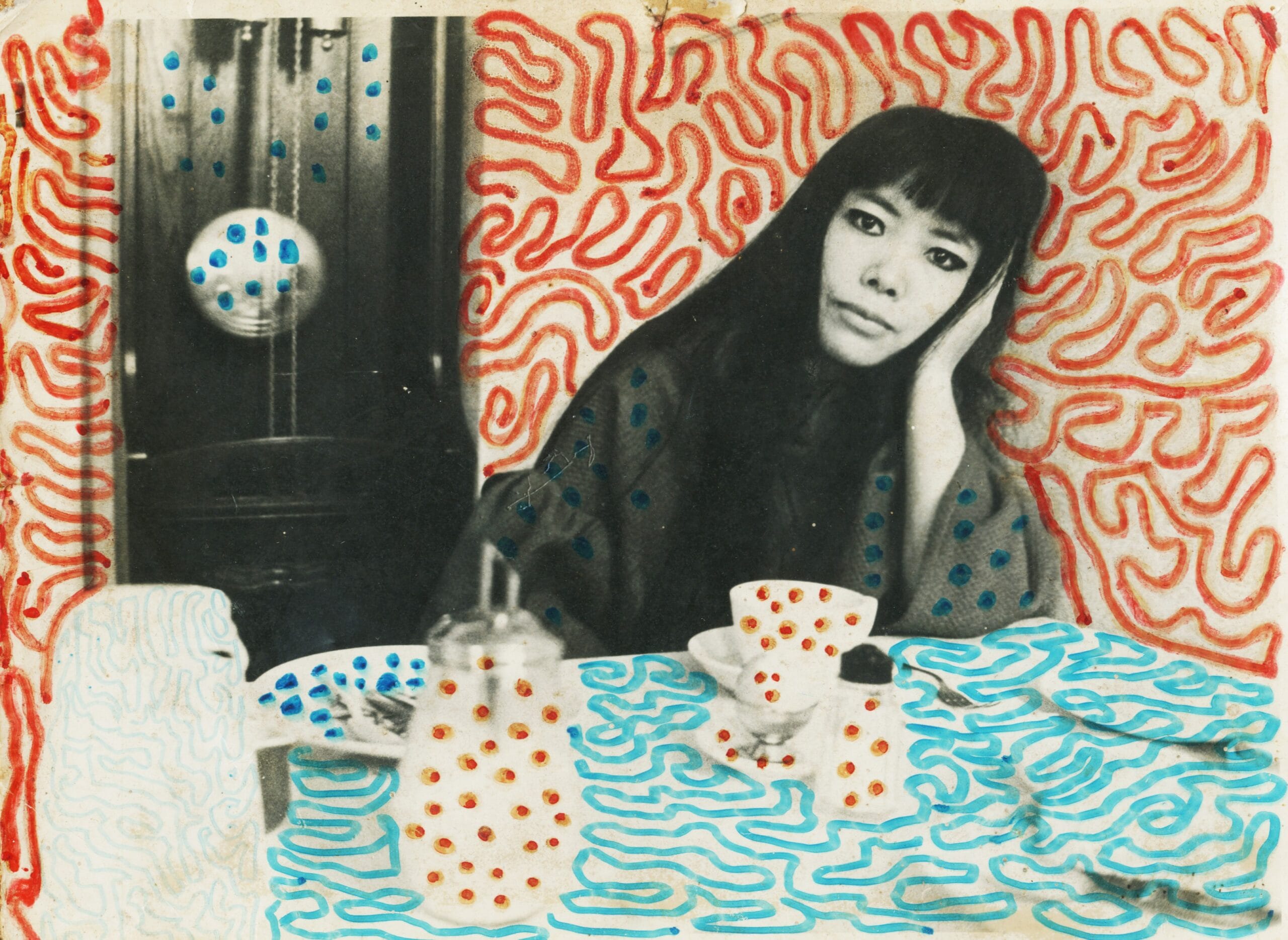 Yayoi Kusama zittend aan een tafel, bewerkt met haar kenmerkende kleurrijke stippen en golvende patronen
