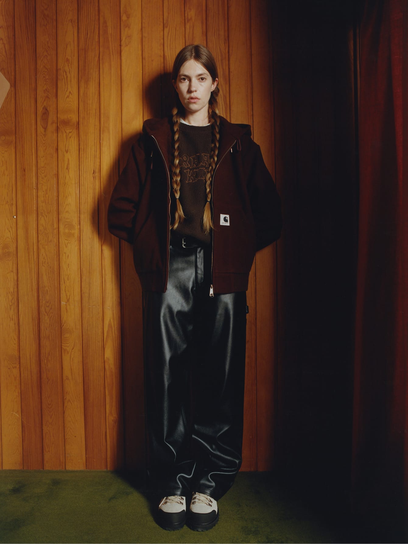 Vrouw in een donkerbruin Carhartt WIP jack, een bruine trui en een zwarte broek van kunstleer tegen een houten wand