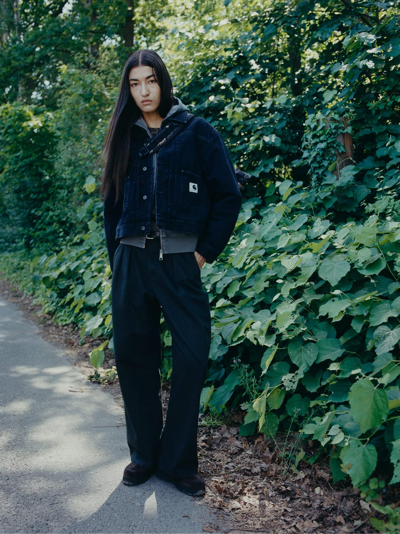 Model in een donkerblauw OG Trucker Jacket van denim, een grijze hoodie en een wijde zwarte broek voor een groene heg