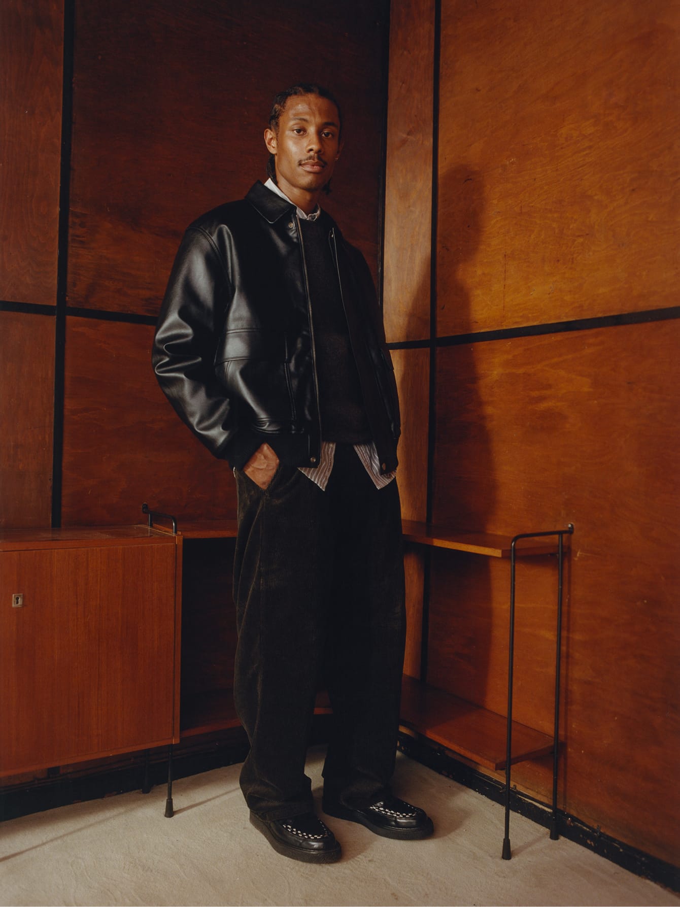 Man in een zwart kunstleren Arizona Jacket, een donkere trui en een wijde ribfluwelen broek tegen een houten wand