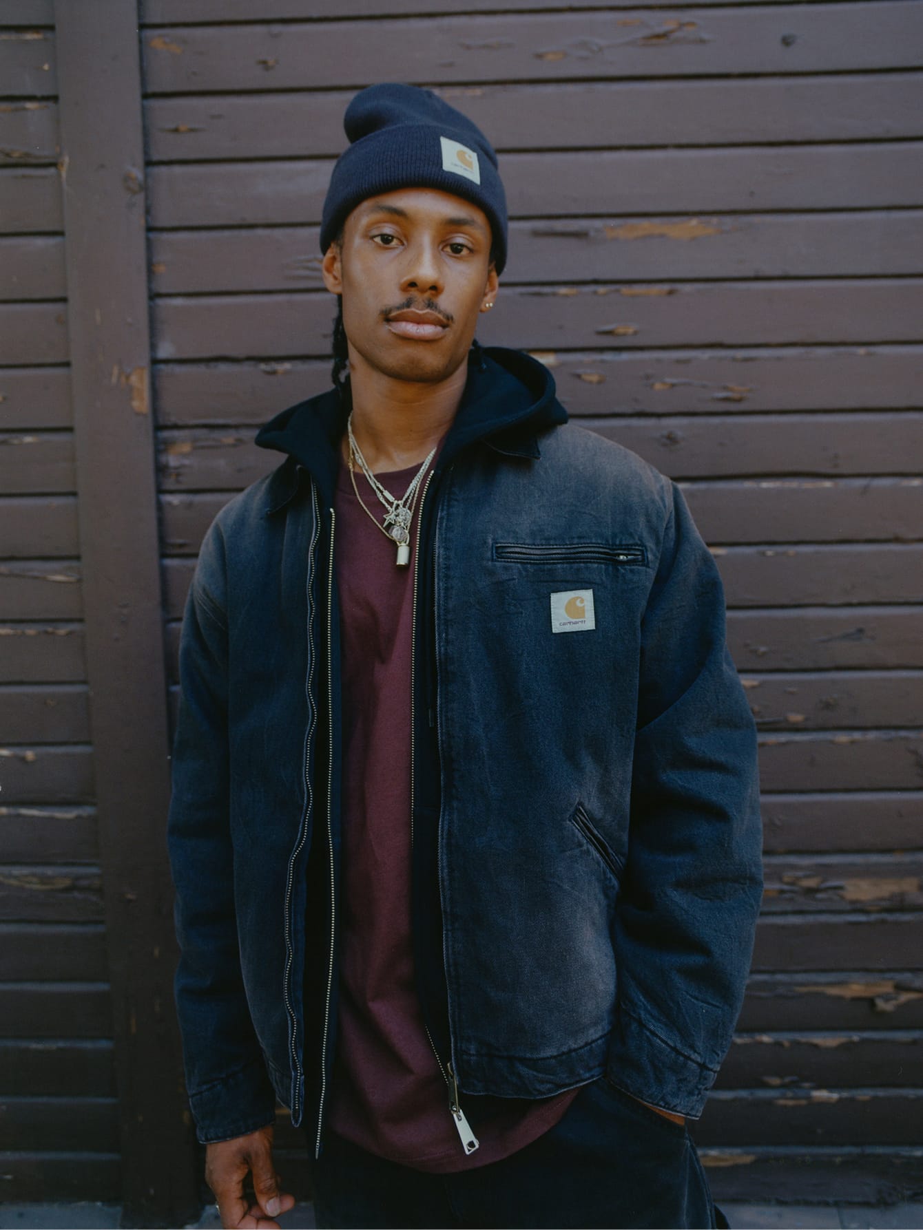 Man in een donkerblauw gewassen OG Detroit Jacket met een zwarte hoodie, bordeauxrood shirt en een donkerblauwe muts