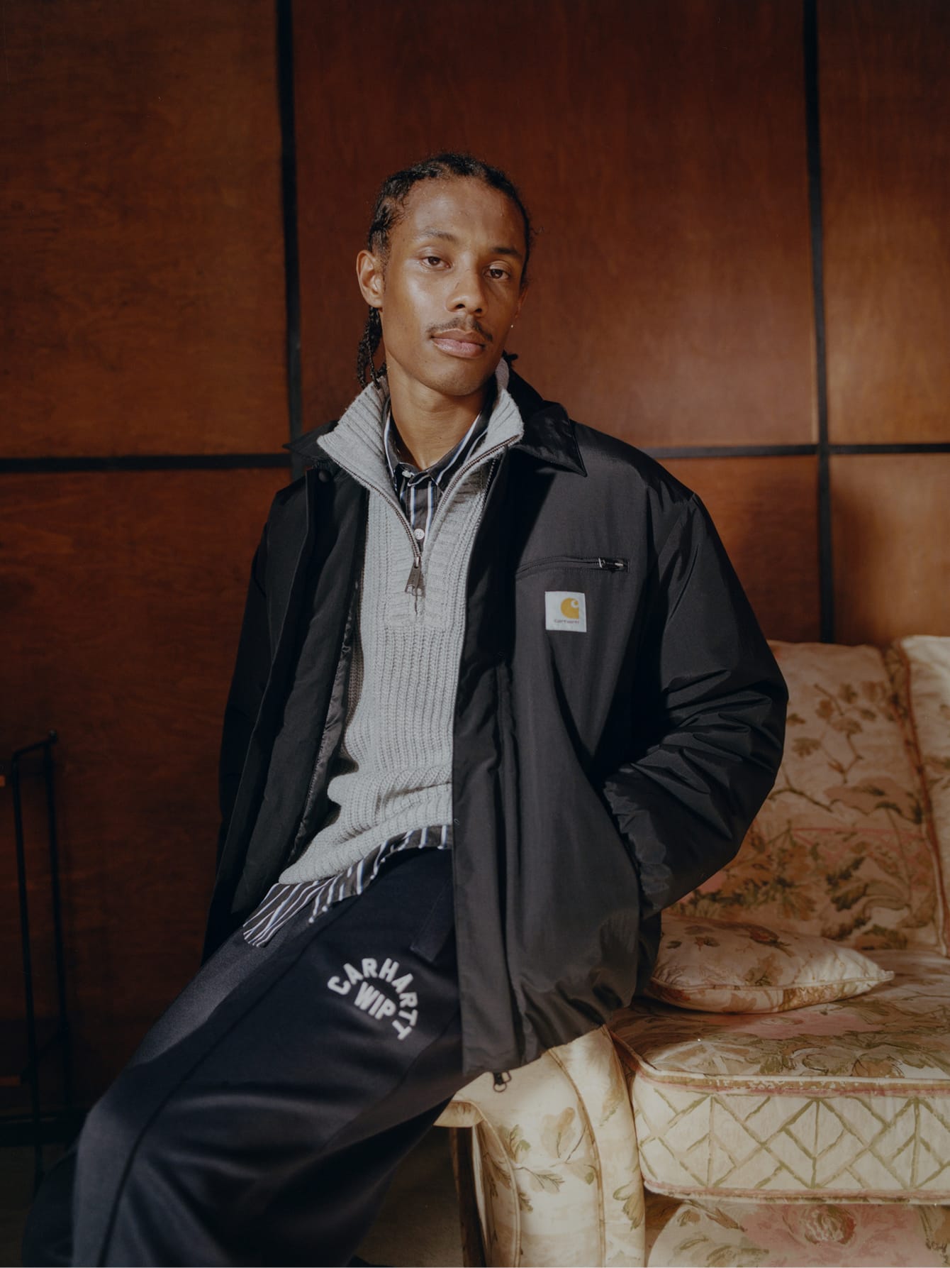Model draagt een zwart Carhartt WIP jack over een grijze gebreide trui met rits en een donkere broek met logo