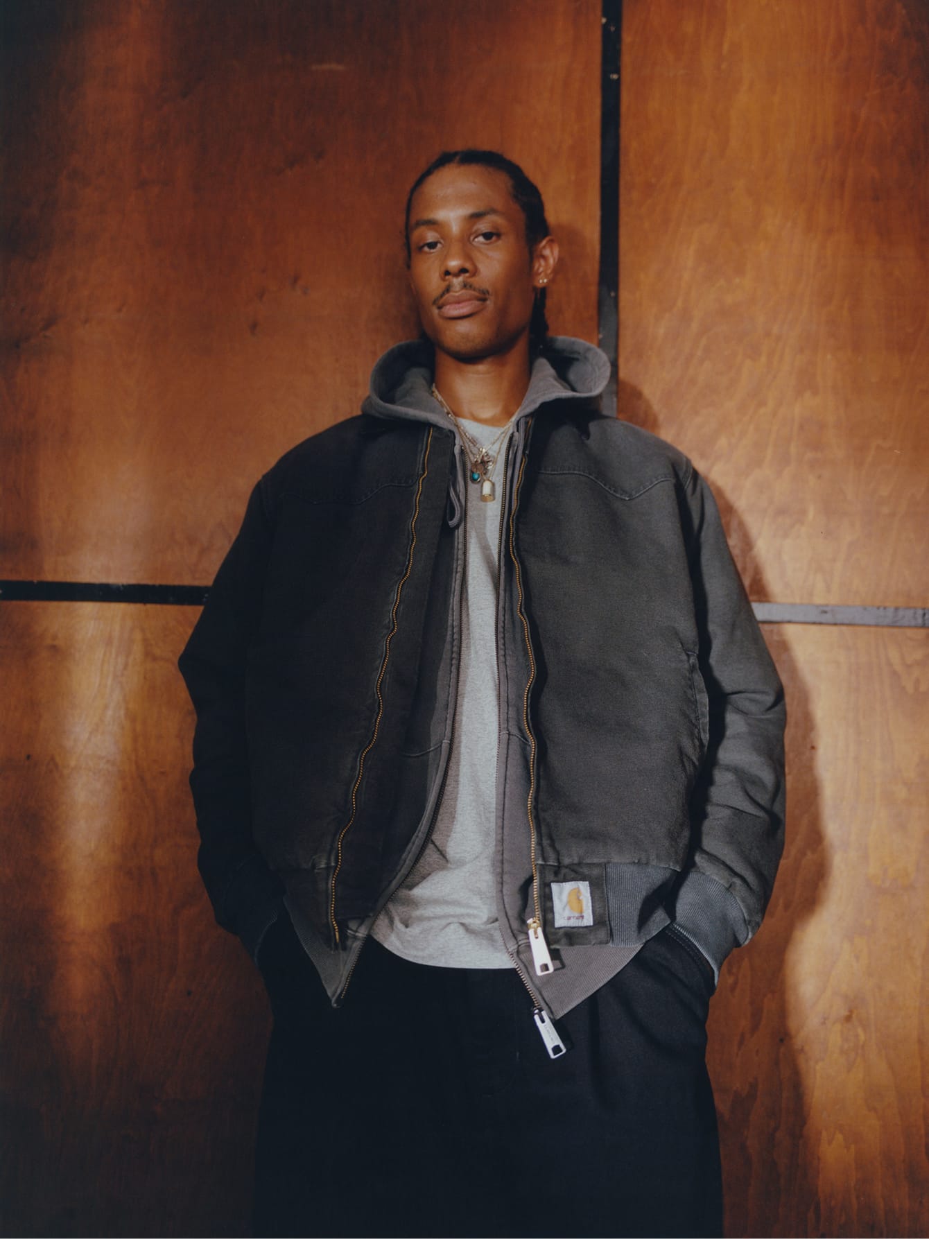 Man in een donkergrijs Carhartt WIP-jack met rits over een grijze hoodie en T-shirt tegen een houten achtergrond