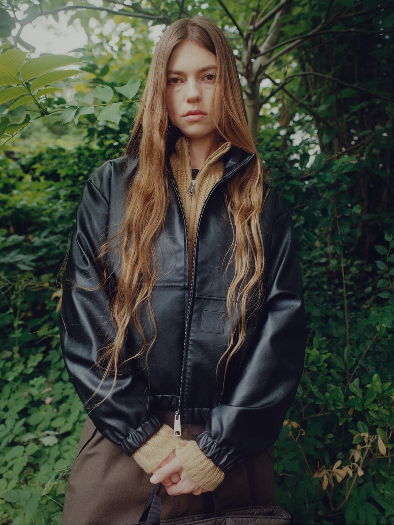 Vrouw in een zwart kunstleren Arizona Jacket over een beige gebreide trui, staand voor een groene achtergrond