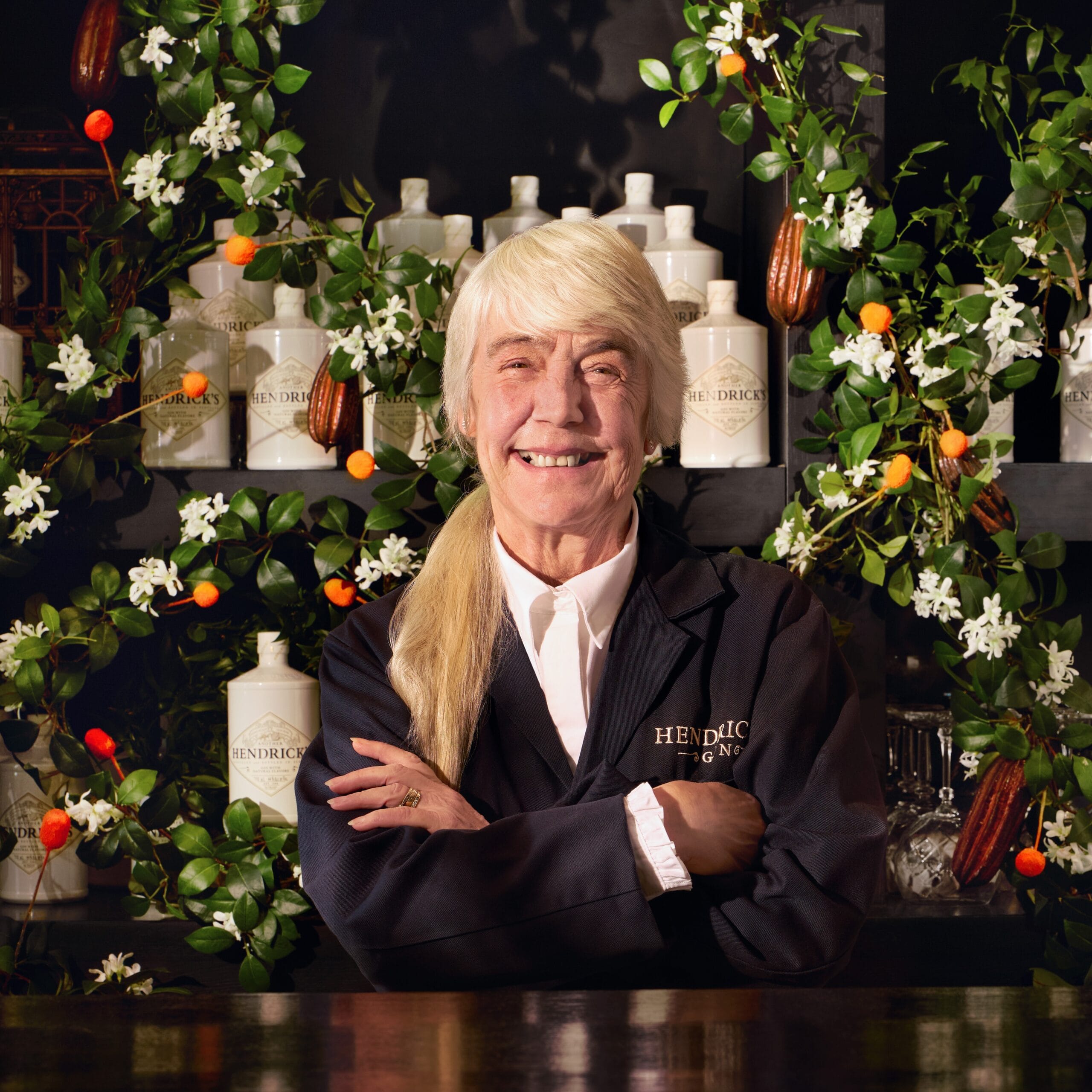 Portret van Master Distiller Lesley Gracie voor een wand met witte flessen Another Hendrick’s gin, bloemen en cacaovruchten