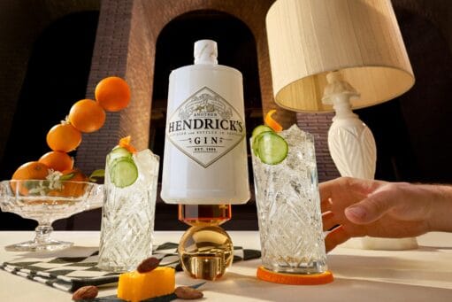Een witte fles Hendrick's Gin omringd door twee glazen gin-tonic met komkommer en een schaal met sinaasappels