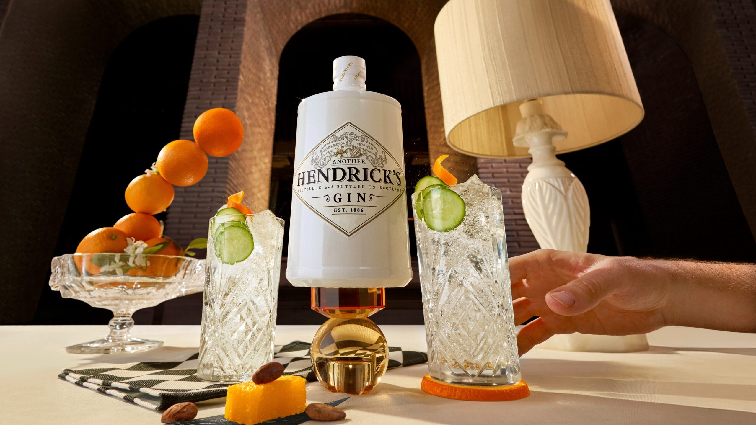 Een witte fles Hendrick's Gin omringd door twee glazen gin-tonic met komkommer en een schaal met sinaasappels