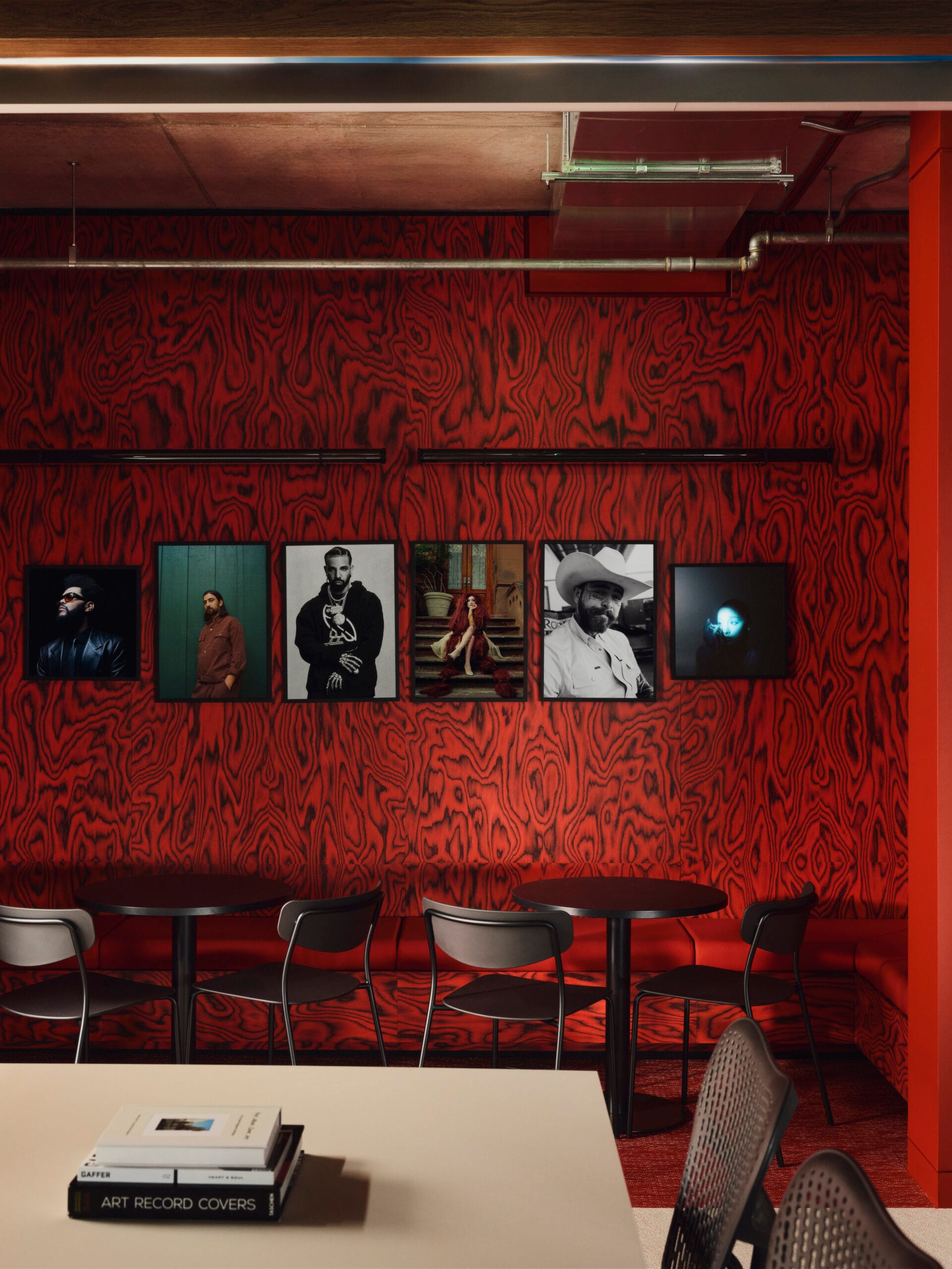 Een wand met een rood-zwart houtnerfpatroon waaraan portretten van artiesten hangen boven een zithoek met zwarte tafels