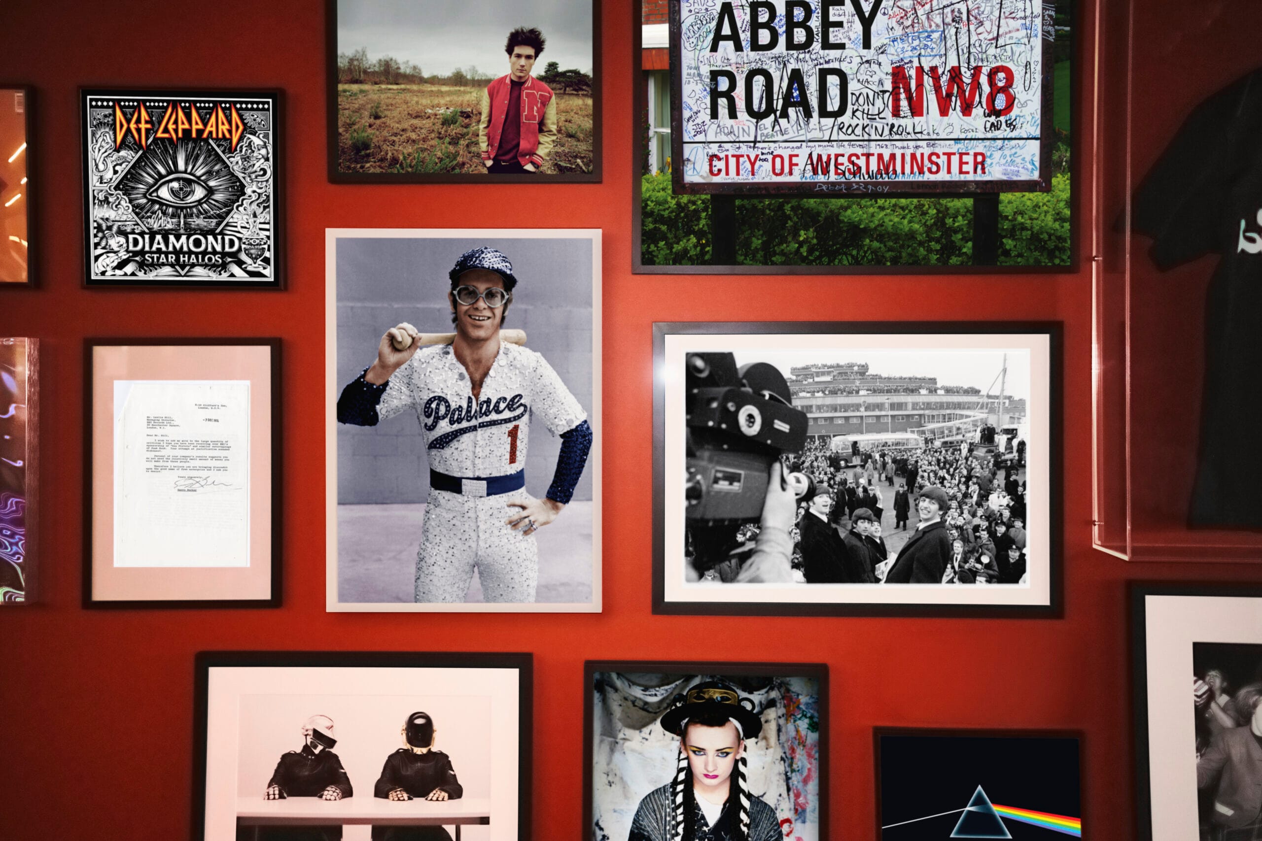 Een rode wand met ingelijste muziekmemorabilia, waaronder foto's van Elton John, The Beatles, Boy George en Daft Punk