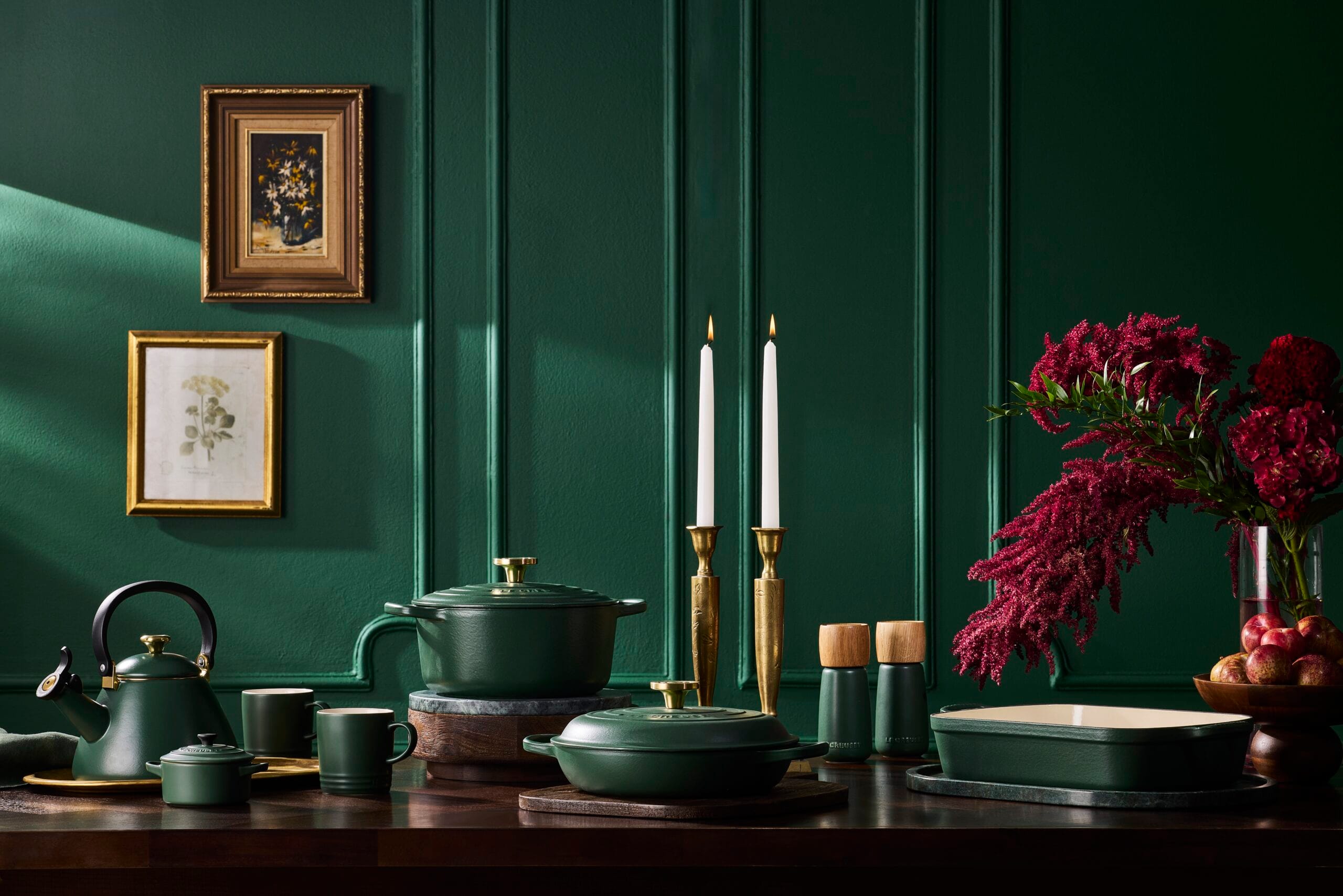 Donkergroene collectie pannen, fluitketel, mokken en ovenschalen op een houten tafel voor een groene wand, allemaal in het teken van de nieuwe kleur Foret van Le Creuset