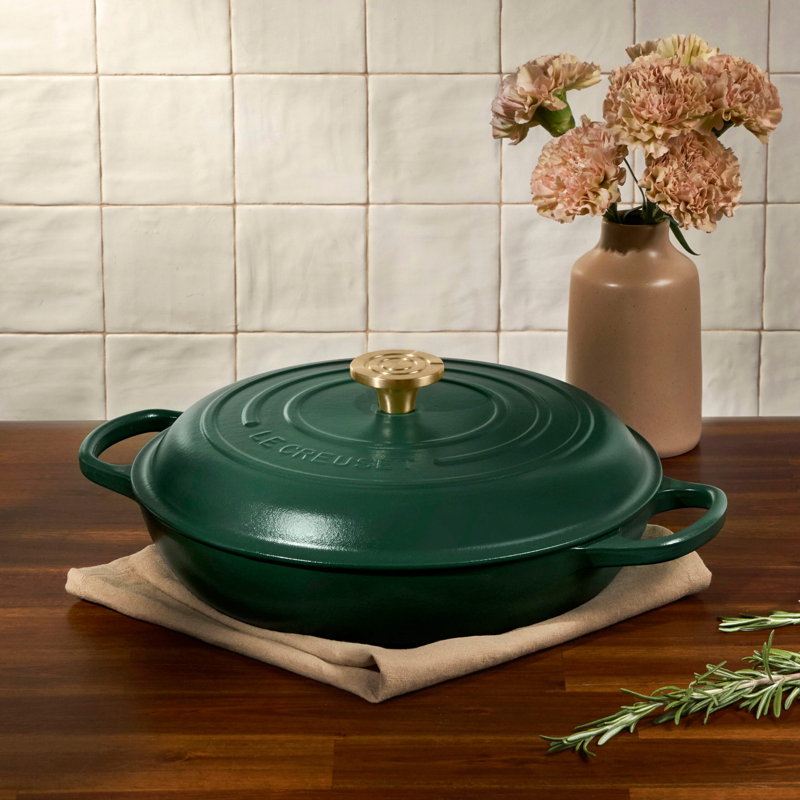 Donkergroene Le Creuset gietijzeren braadpan met gouden knop op een houten tafel naast een vaas bloemen