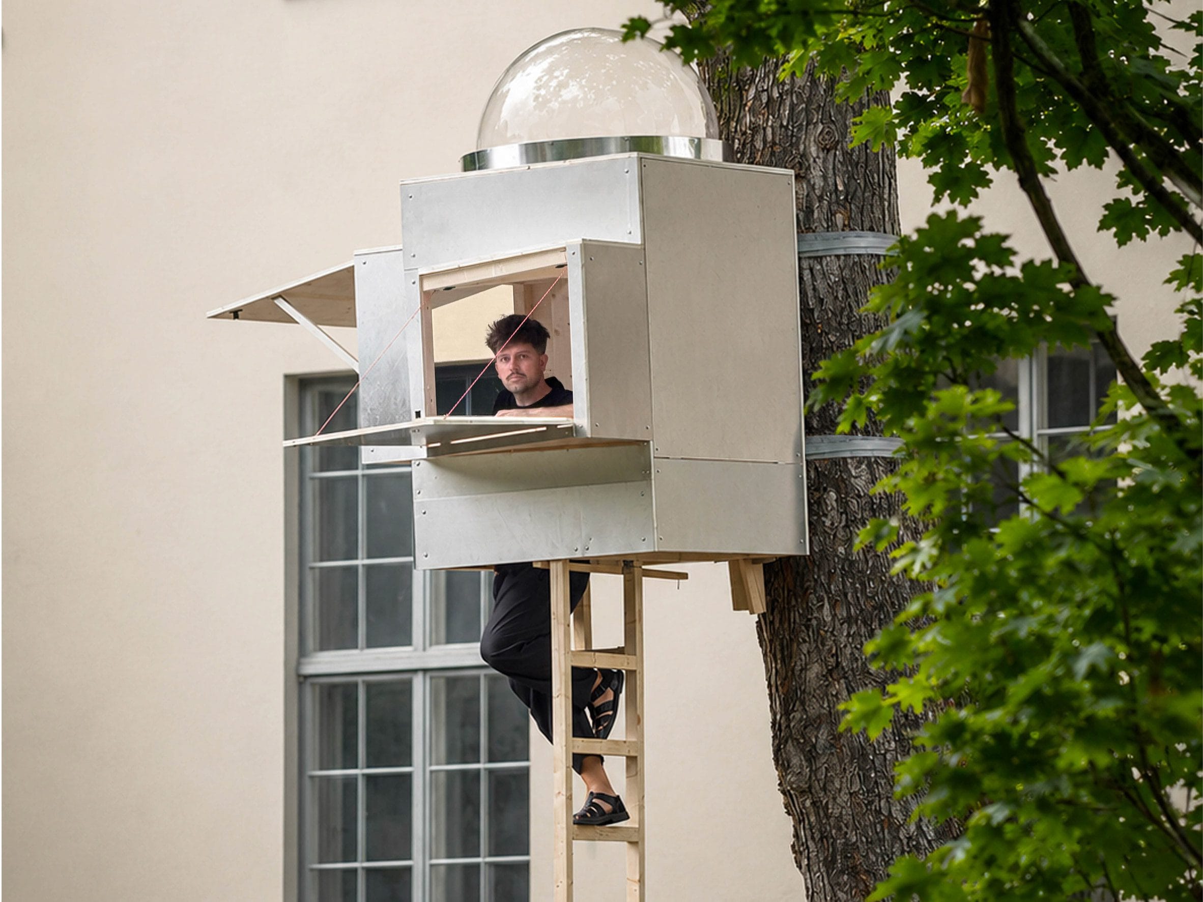 Benedikt Hartl kijkt uit het compacte, aluminiumkleurige Baumhaus Office dat met spanbanden aan een boom is bevestigd