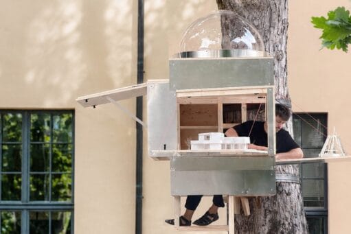 Man zit in een compacte houten en metalen boomhut met opengeklapte luiken waarin maquettes staan