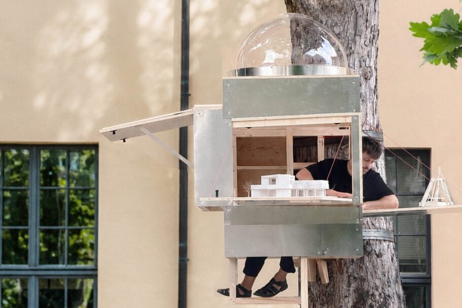 Man zit in een compacte houten en metalen boomhut met opengeklapte luiken waarin maquettes staan