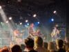 Band treedt live op met zang, viool, gitaar en drums op een verlicht podium voor een publiek