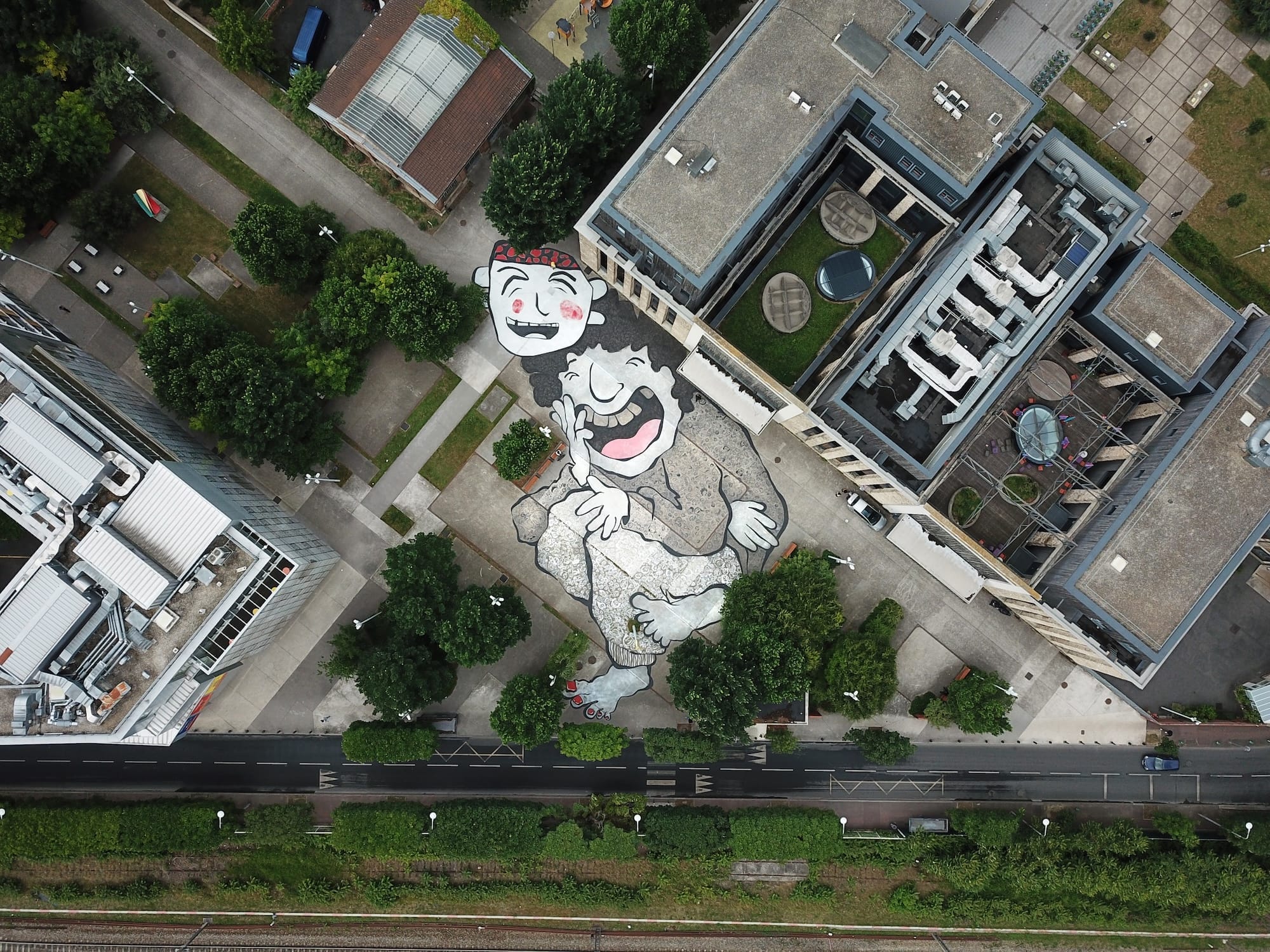 Luchtfoto van een gigantische muurschildering op een plein tussen gebouwen, waarop twee lachende figuren zijn afgebeeld