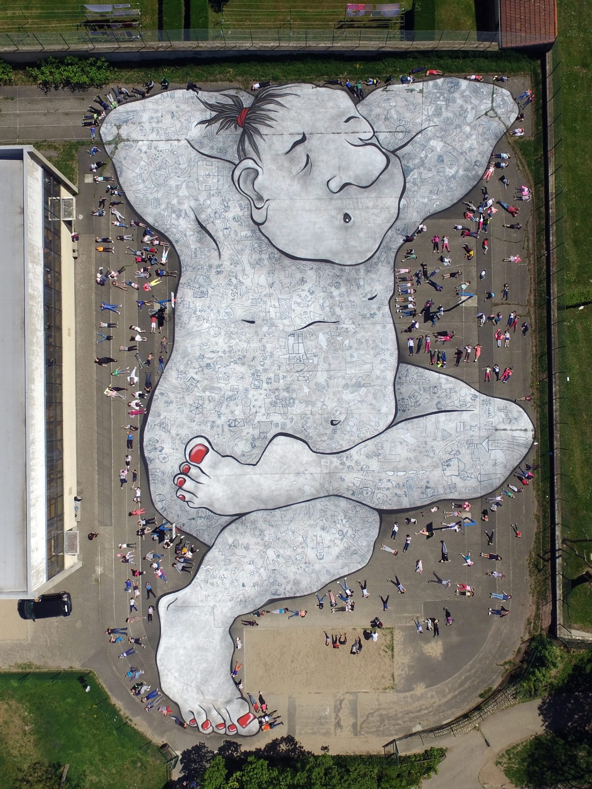 Luchtfoto van een gigantische muurschildering van een slapende reus op een grijs plein, omringd door kleine mensen