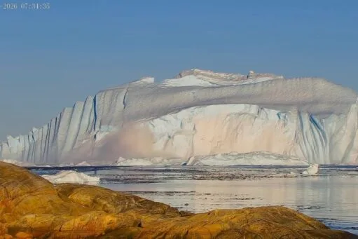 Een enorme ijsberg drijft in het water voor een rotsachtige kust onder een helderblauwe lucht