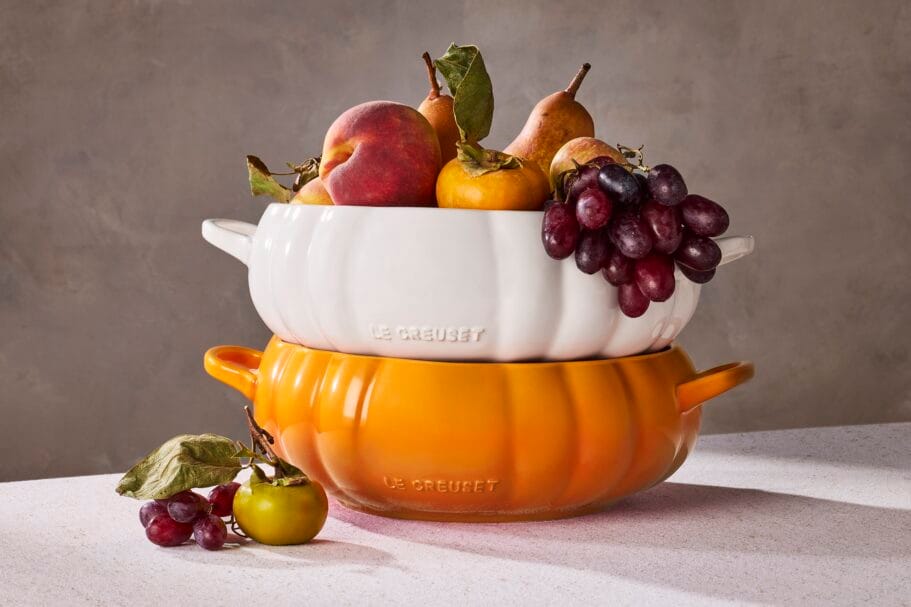 Gestapelde witte en oranje pompoenvormige Le Creuset-schalen gevuld met herfstfruit zoals peren, druiven en perziken