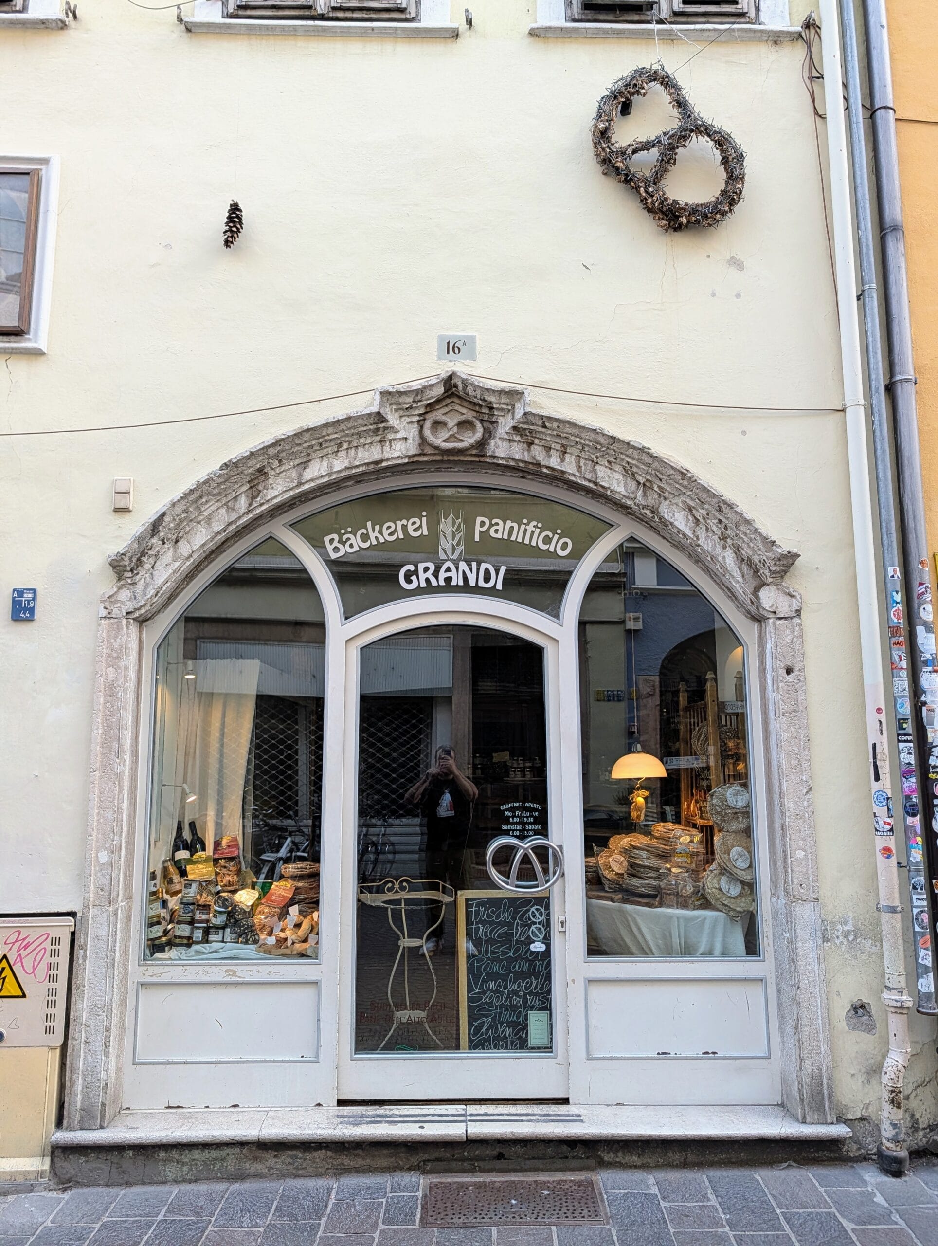 Gevel van bakkerij Bäckerei Panificio Grandi met een stenen boog, krakelingversieringen en broden in de etalage