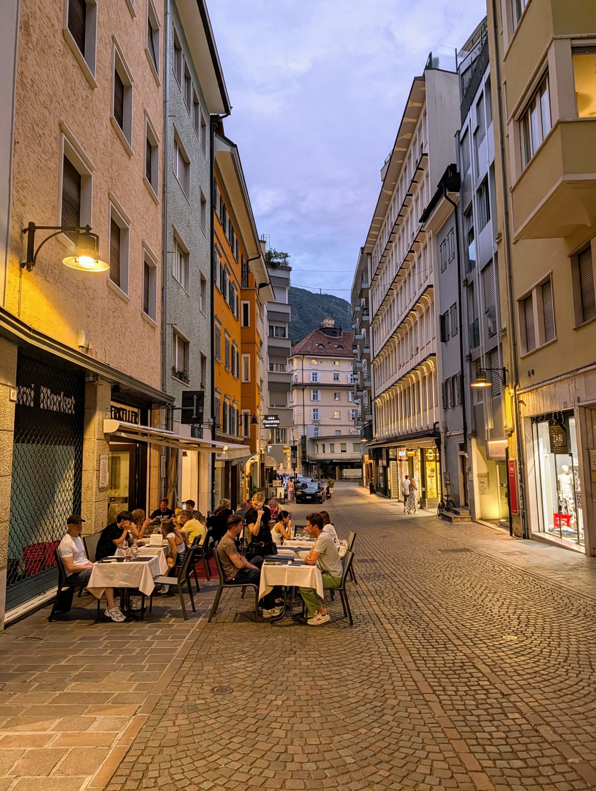Mensen dineren aan gedekte tafeltjes in een sfeervolle, geplaveide straat in Bolzano tijdens de schemering