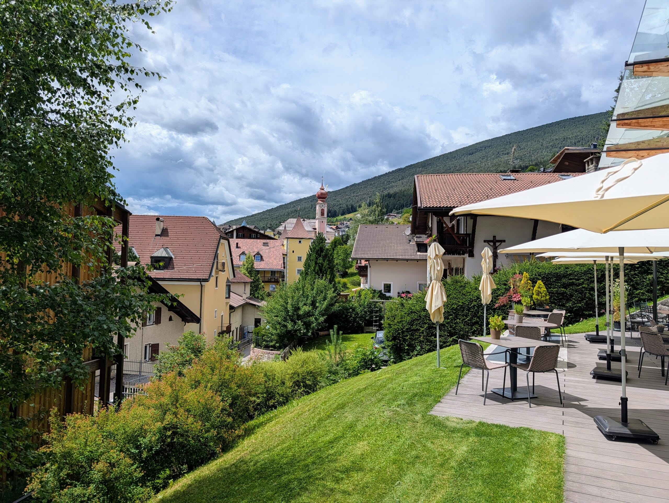 Terras met zitjes en uitzicht op traditionele huizen, een kerktorentje en groene bergen in Zuid-Tirol