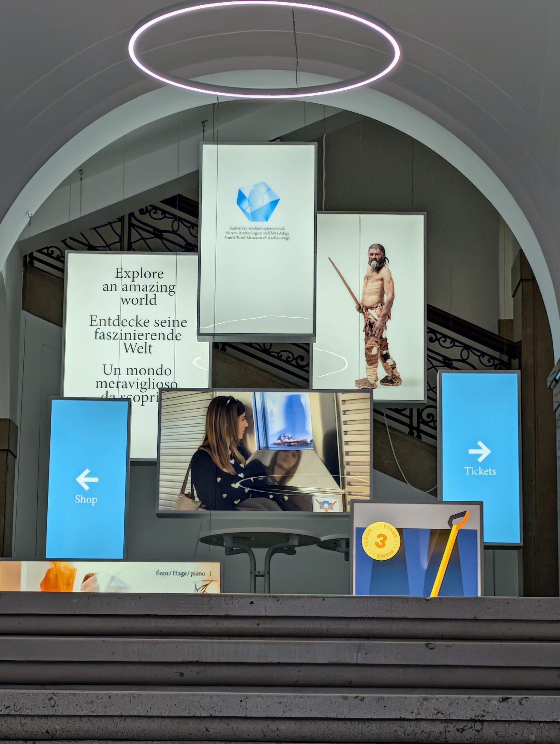 Schermen bij de entree van het Südtiroler Archäologiemuseum in Bolzano met een afbeelding van Ötzi en route-aanwijzingen