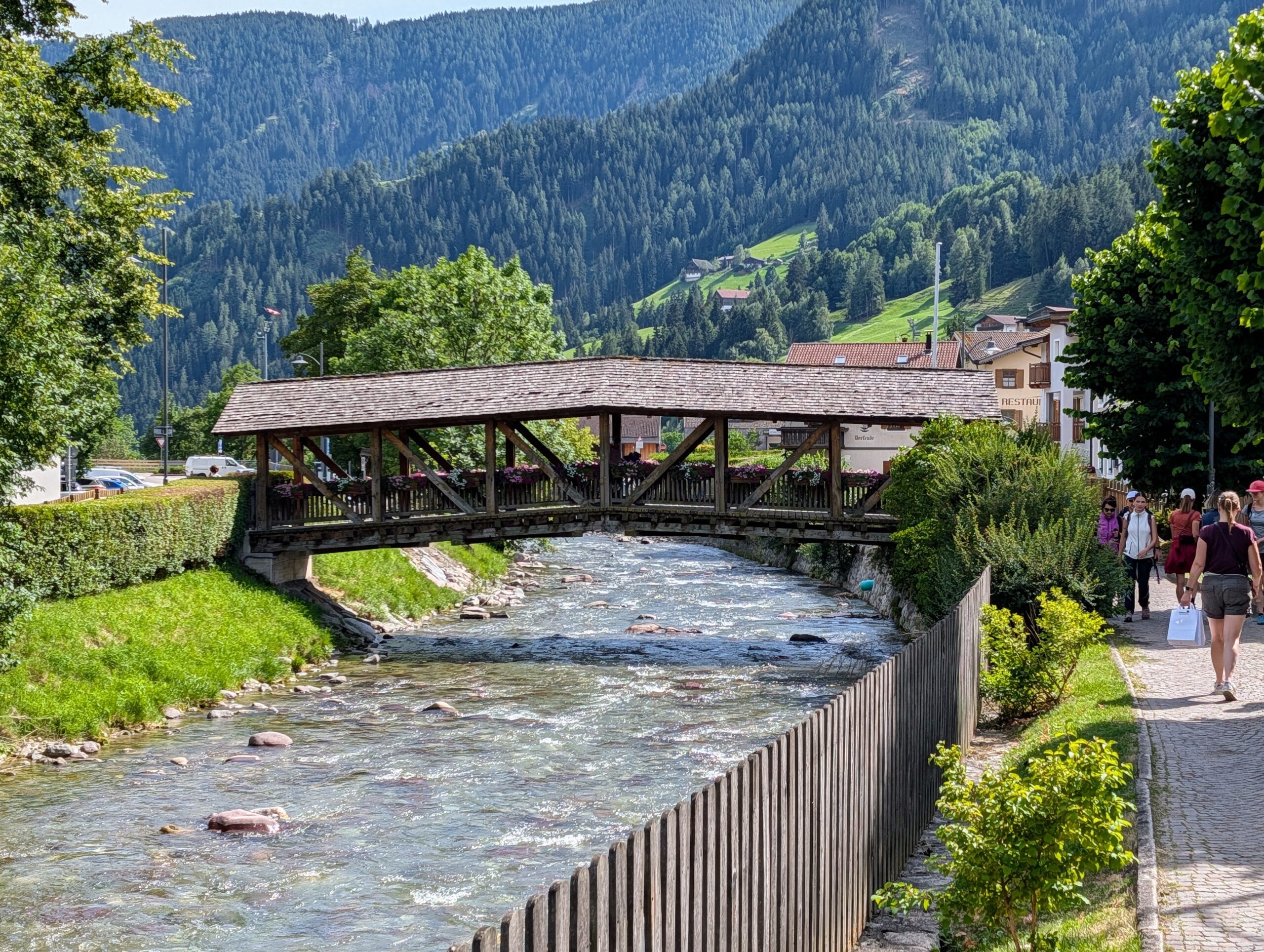 Overdekte houten brug over een kabbelende beek in een bergdorpje, met groene beboste bergen op de achtergrond