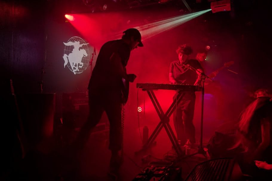Muzikanten treden live op een podium op onder fel rood licht en rook, met een toetsenist en gitarist