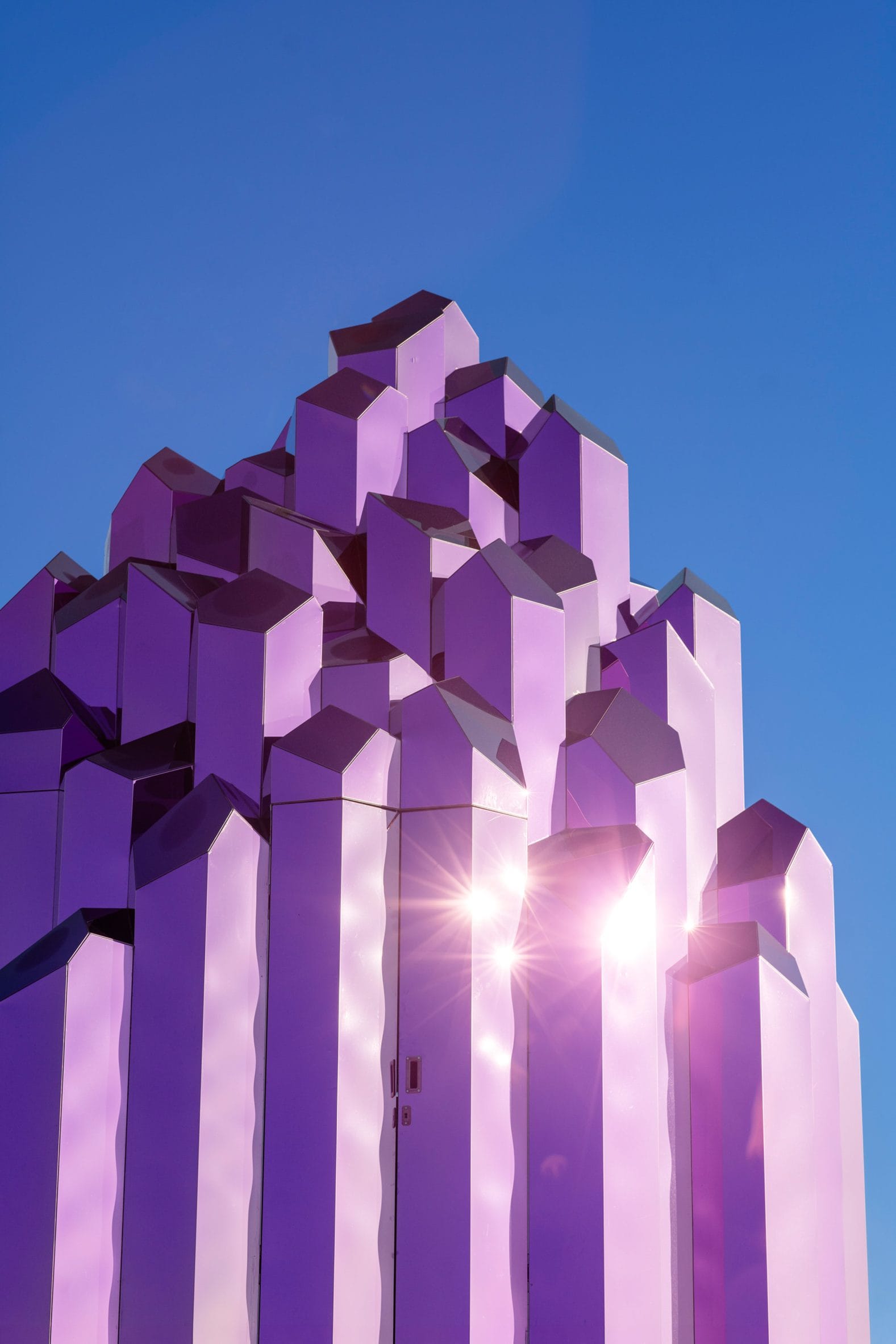 De Lithium Crystal Sauna in Skellefteå: een roze, kristalvormig bouwwerk van spiegelend staal tegen een blauwe lucht
