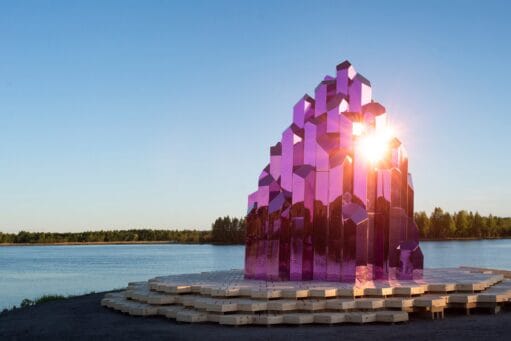 Een grote, paarse kristalvormige sculptuur van spiegels op een houten platform aan de oever van een meer bij zonsondergang