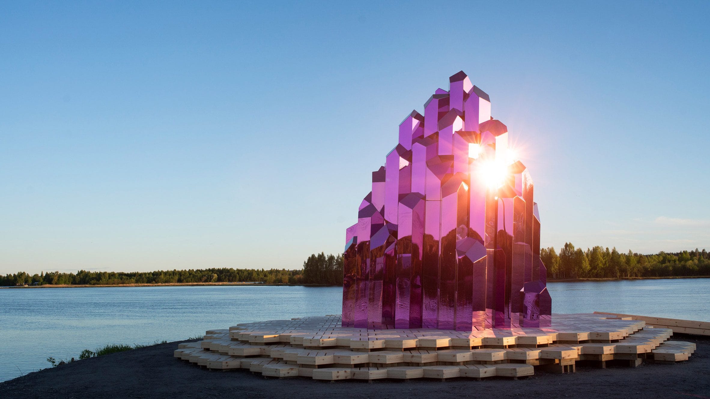 De roze, kristalvormige Lithium Crystal Sauna op een houten platform aan de oever van een rivier in Skellefteå
