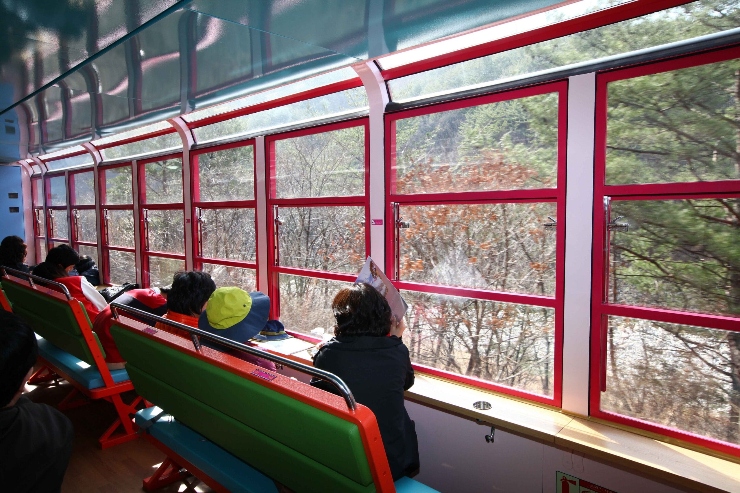 Passagiers in de V-Train kijken door grote panoramaramen met rode kozijnen uit over een bebost berglandschap