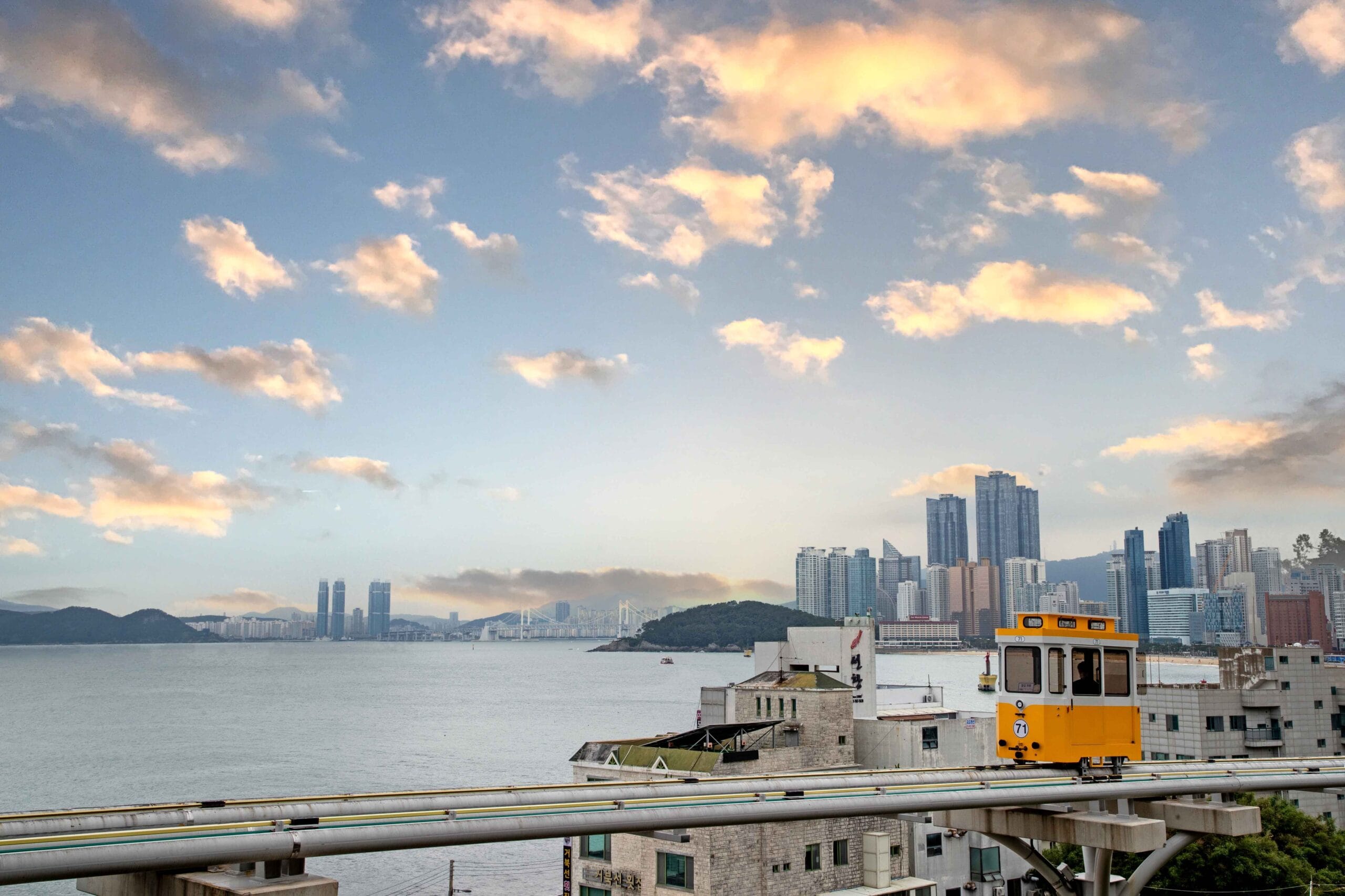 Een gele Busan Sky Capsule op een verhoogd spoor langs de kustlijn van Haeundae met de skyline van de stad op de achtergrond