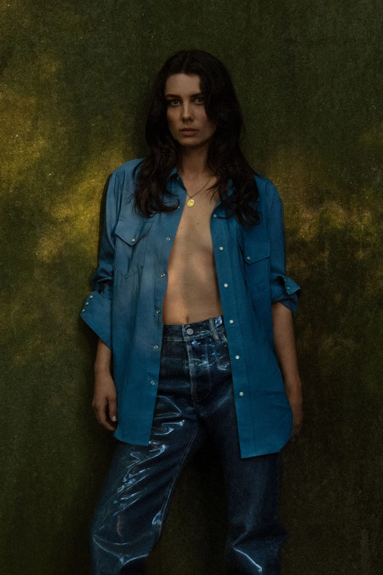 Portret van muzikant met een openvallend blauw denim overhemd en glanzende jeans tegen een donkere achtergrond