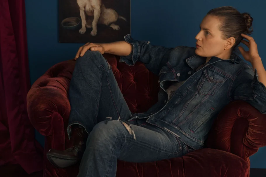 Persoon in spijkerjas en jeans zit ontspannen achterover in een bordeauxrode fluwelen fauteuil tegen een blauwe muur