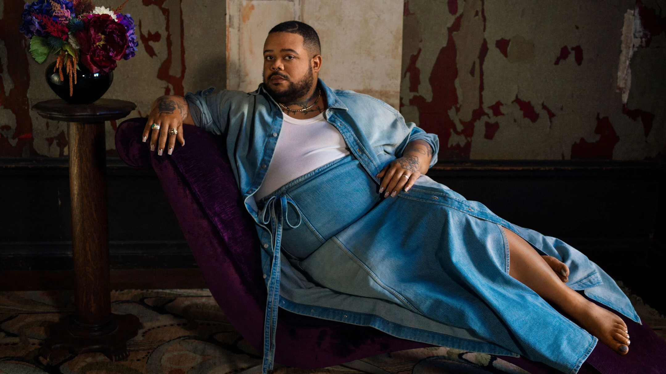 Jonathan Lyndon Chase liggend op een paarse chaise longue in een denim rok, open spijkeroverhemd en wit hemd
