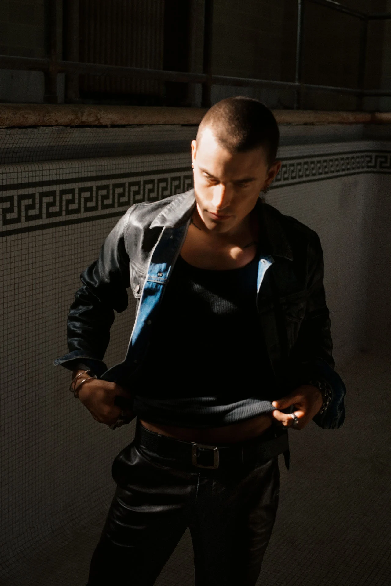 Jordan Hemingway poseert in een donker denim jack, zwarte tanktop en leren broek in een leeg betegeld zwembad