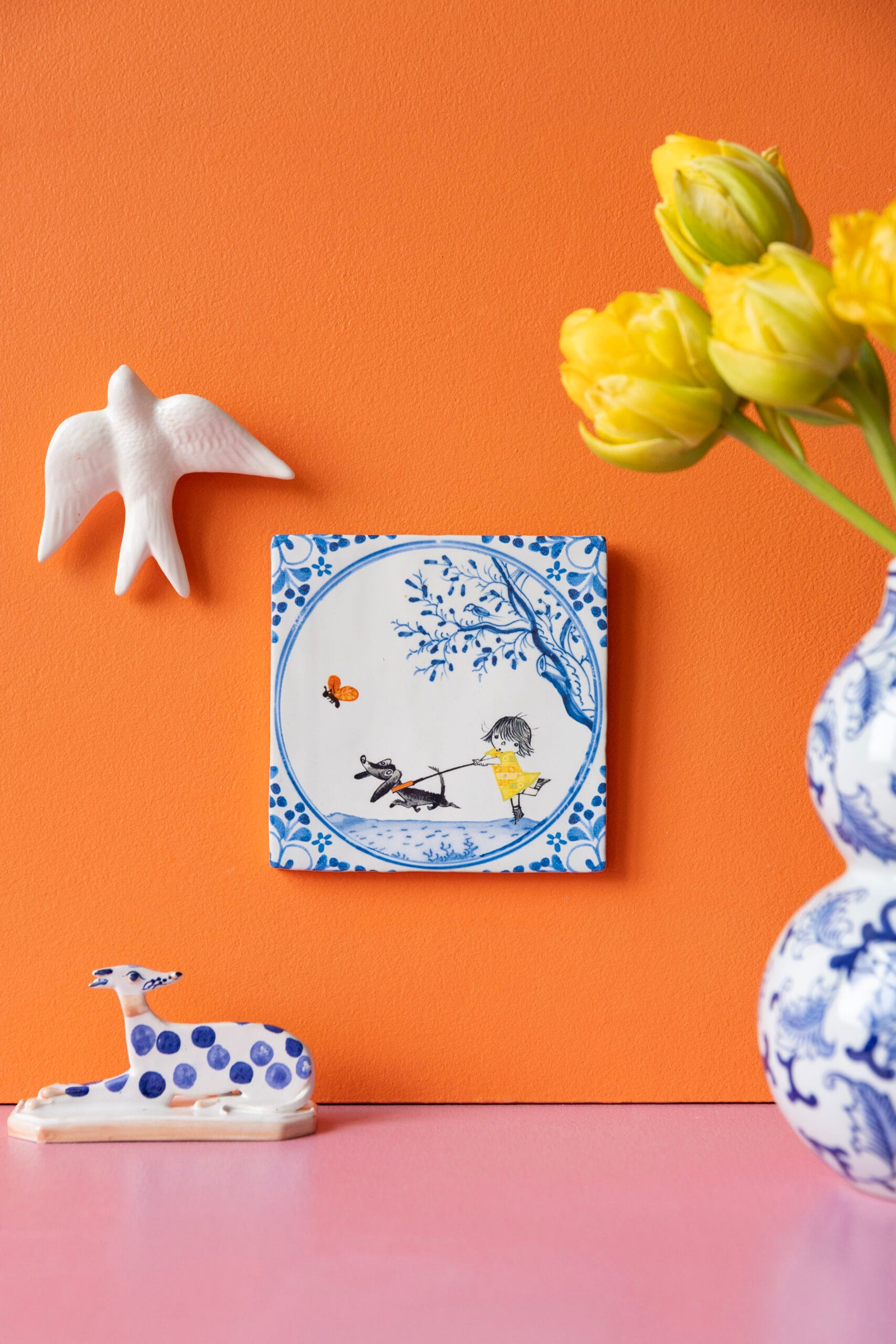 Delfts blauwe tegel genaamd Beste Vriendjes met een meisje in een geel jurkje dat een hondje uitlaat tegen een oranje muur met decoraties, Uit de collectie van StoryTiles x Fiep Westendorp
