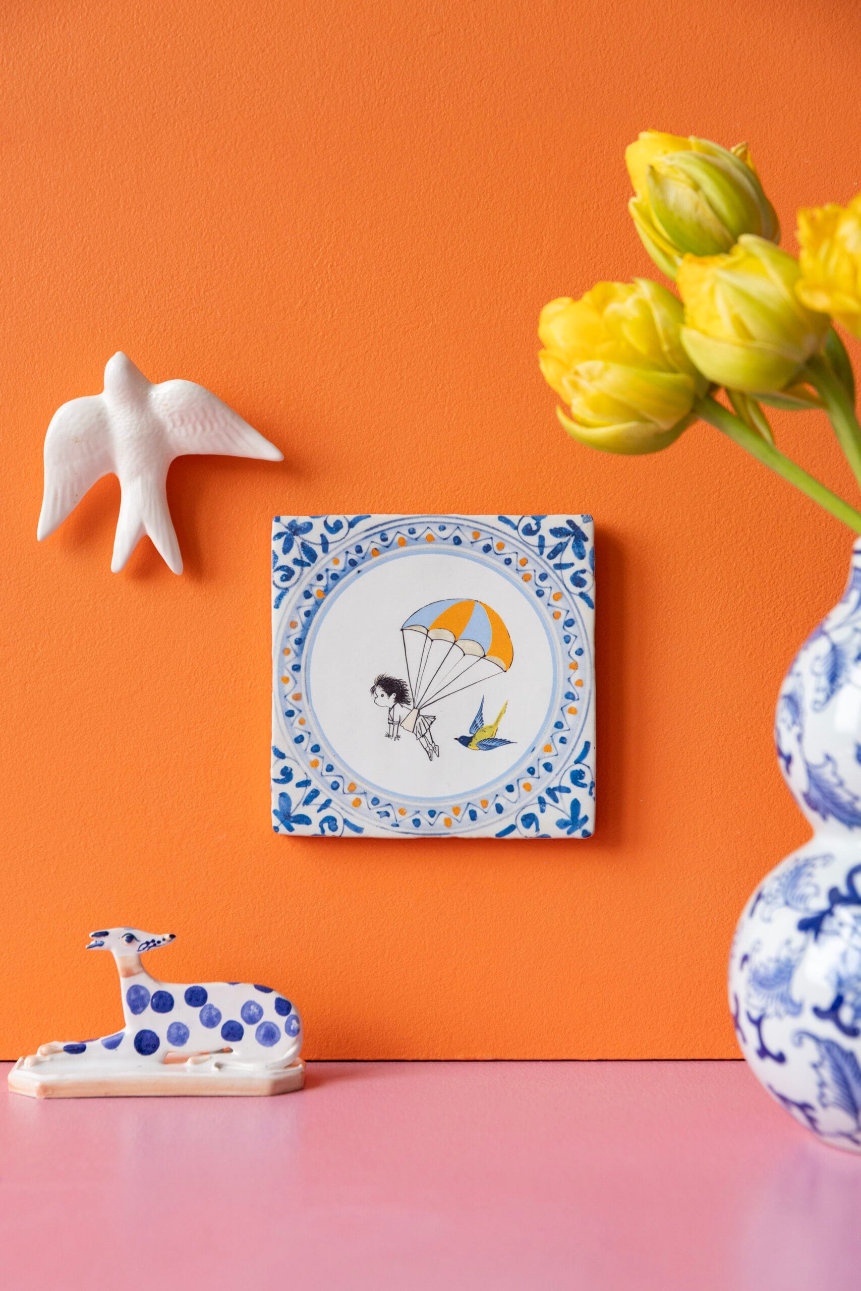 Keramische tegel genaamd Droomvlucht met een illustratie van een parachutespringer aan een oranje wand, omringd door decoratieve beeldjes en gele tulpen, Uit de collectie van StoryTiles x Fiep Westendorp