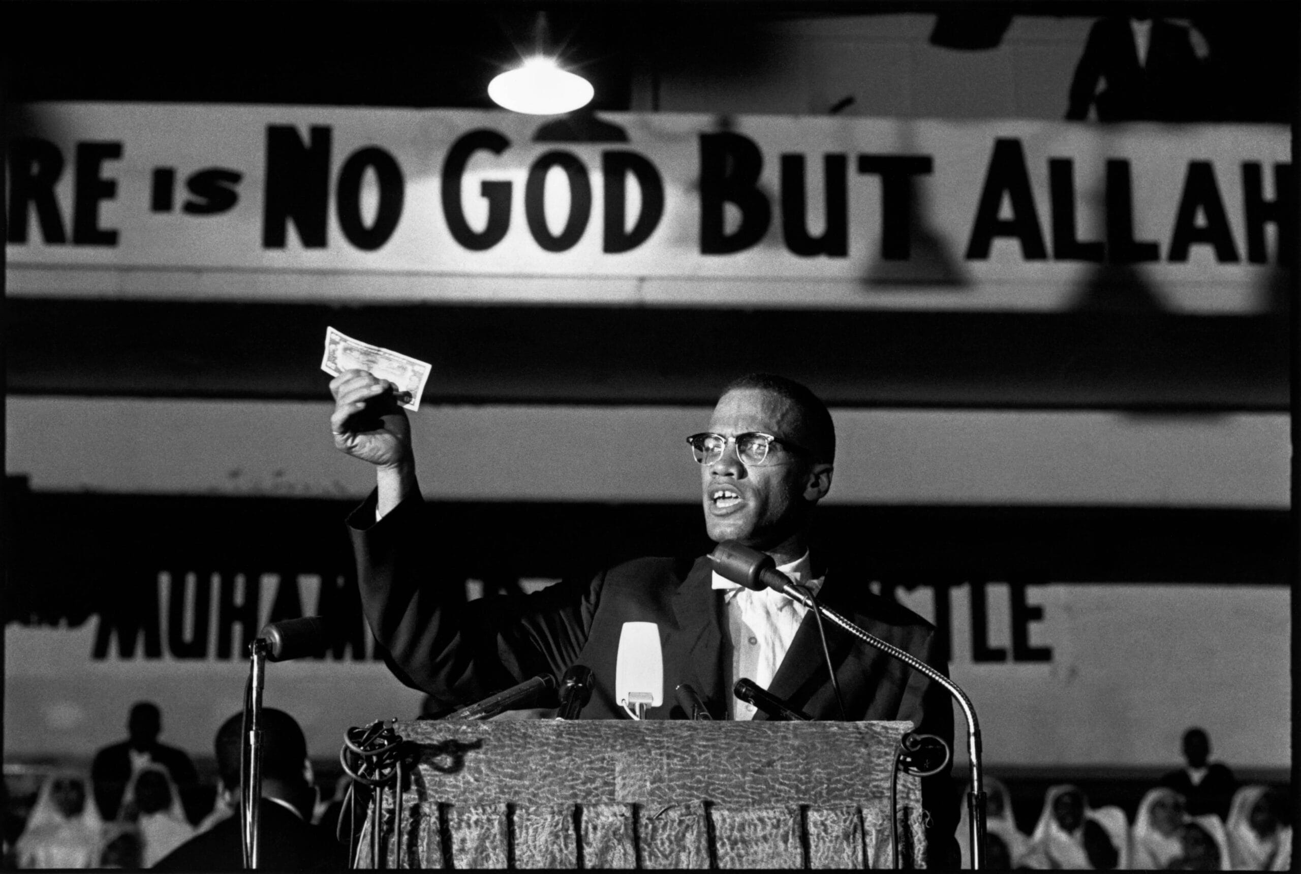 Malcolm X spreekt achter een podium met een dollarbiljet in zijn hand, voor een spandoek met 'There is no God but Allah, gefotografeerd door Eve Arnold in 1961