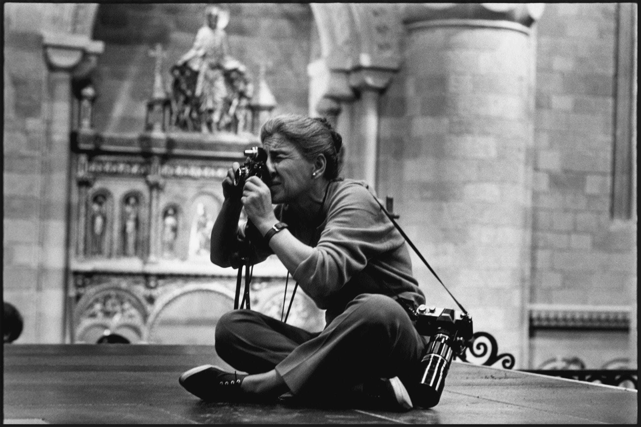 Fotojournalist Eve Arnold in kleermakerszit fotografeert met een analoge camera in een kerk, met een tweede camera over haar schouder