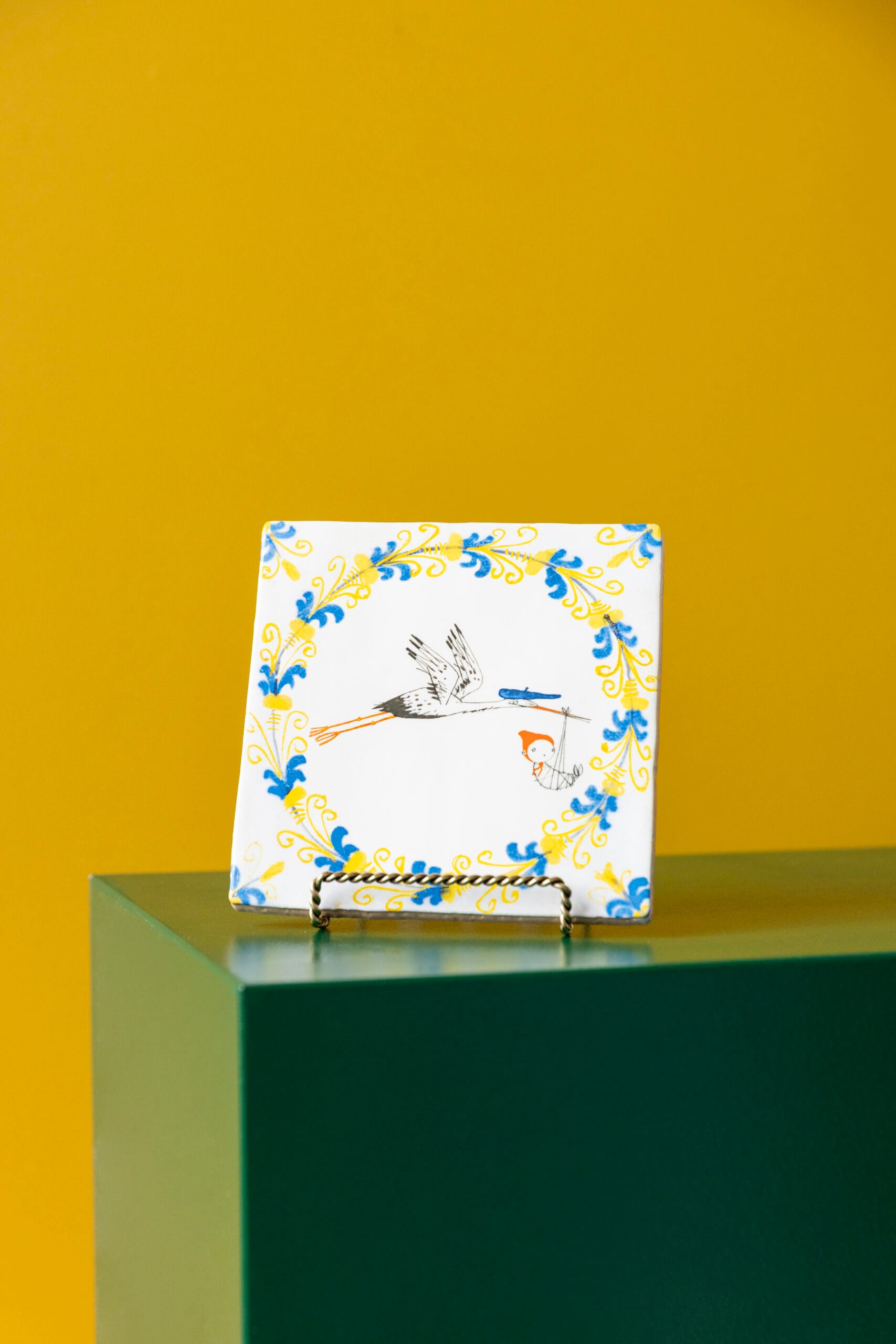 Vierkante tegel genaamd Hallo Kleintje met een illustratie van een vliegende ooievaar die een baby draagt, omringd door een geel-blauwe krans, Uit de collectie van StoryTiles x Fiep Westendorp