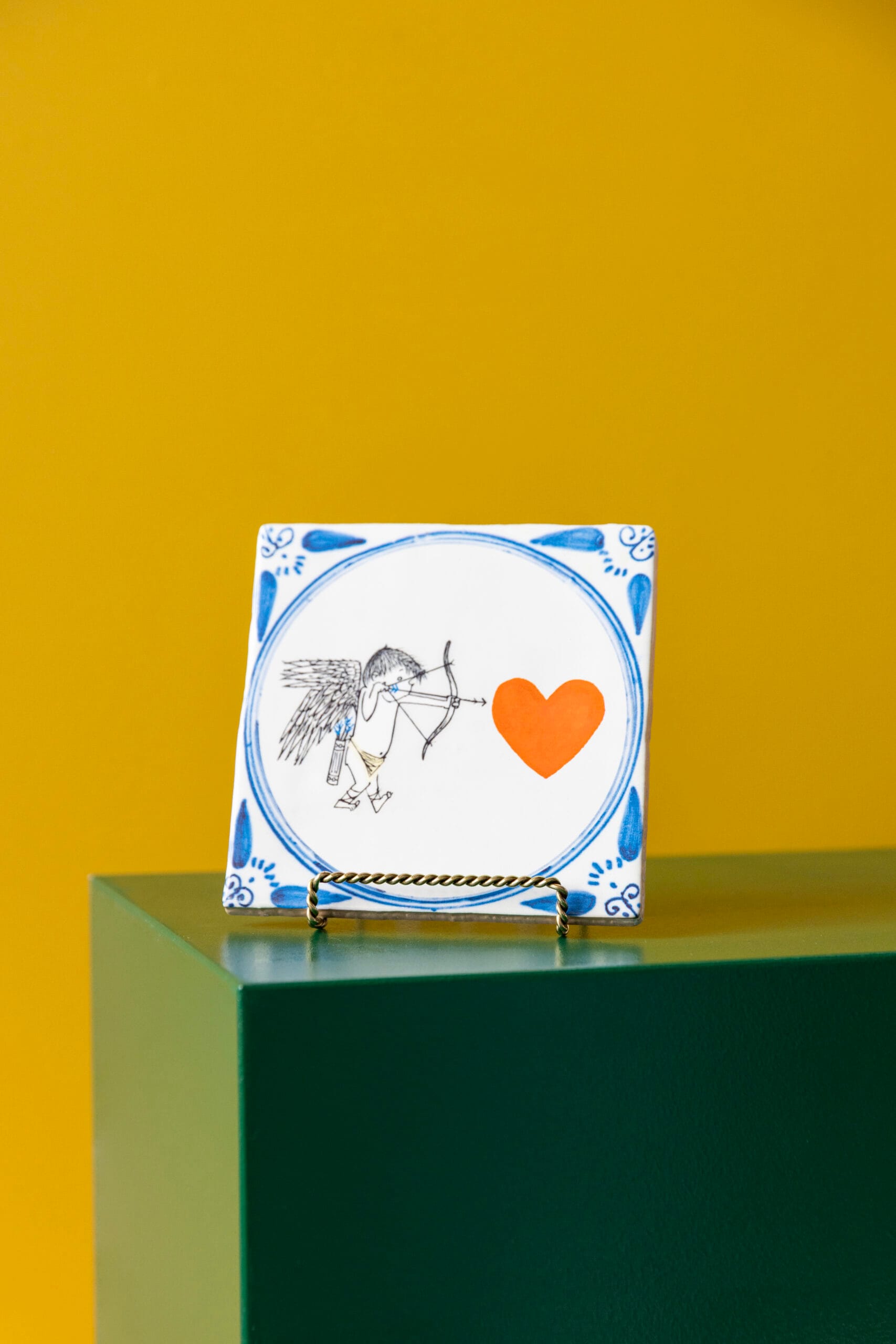 Delftsblauwe tegel genaamd Hartenjager op een standaard met een illustratie van Cupido die met pijl en boog op een oranje hart richt, Uit de collectie van StoryTiles x Fiep Westendorp