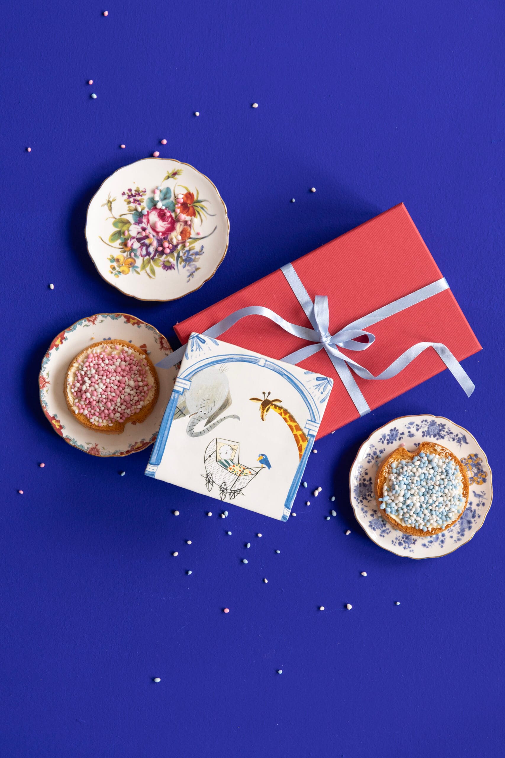 Beschuit met roze en blauwe muisjes op schoteltjes, naast een rood cadeau en een tegel genaamd Jou Bewonderen met een geboorte-illustratie op een blauwe achtergrond. Uit de collectie van StoryTiles x Fiep Westendorp