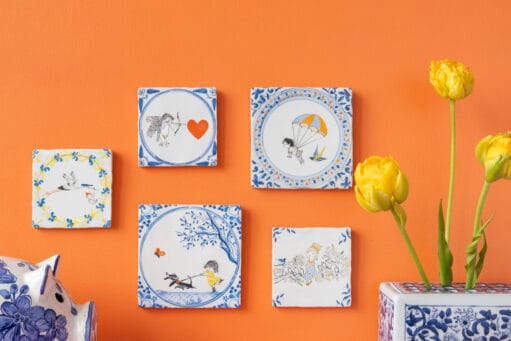 Geïllustreerde Delfts blauwe tegeltjes aan een oranje muur met een vaas gele tulpen en een Delfts blauw spaarvarken, ter promotie van de collectie StoryTiles x Fiep Westendorp