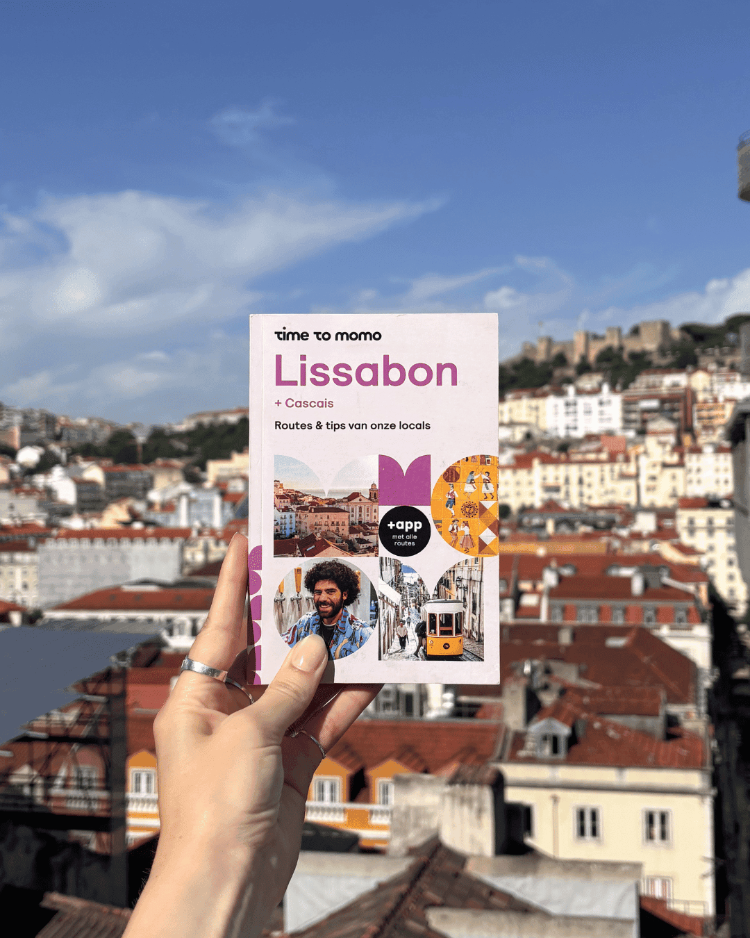 Een hand houdt de Time to Momo-reisgids voor Lissabon vast tegen het uitzicht over de daken van de stad