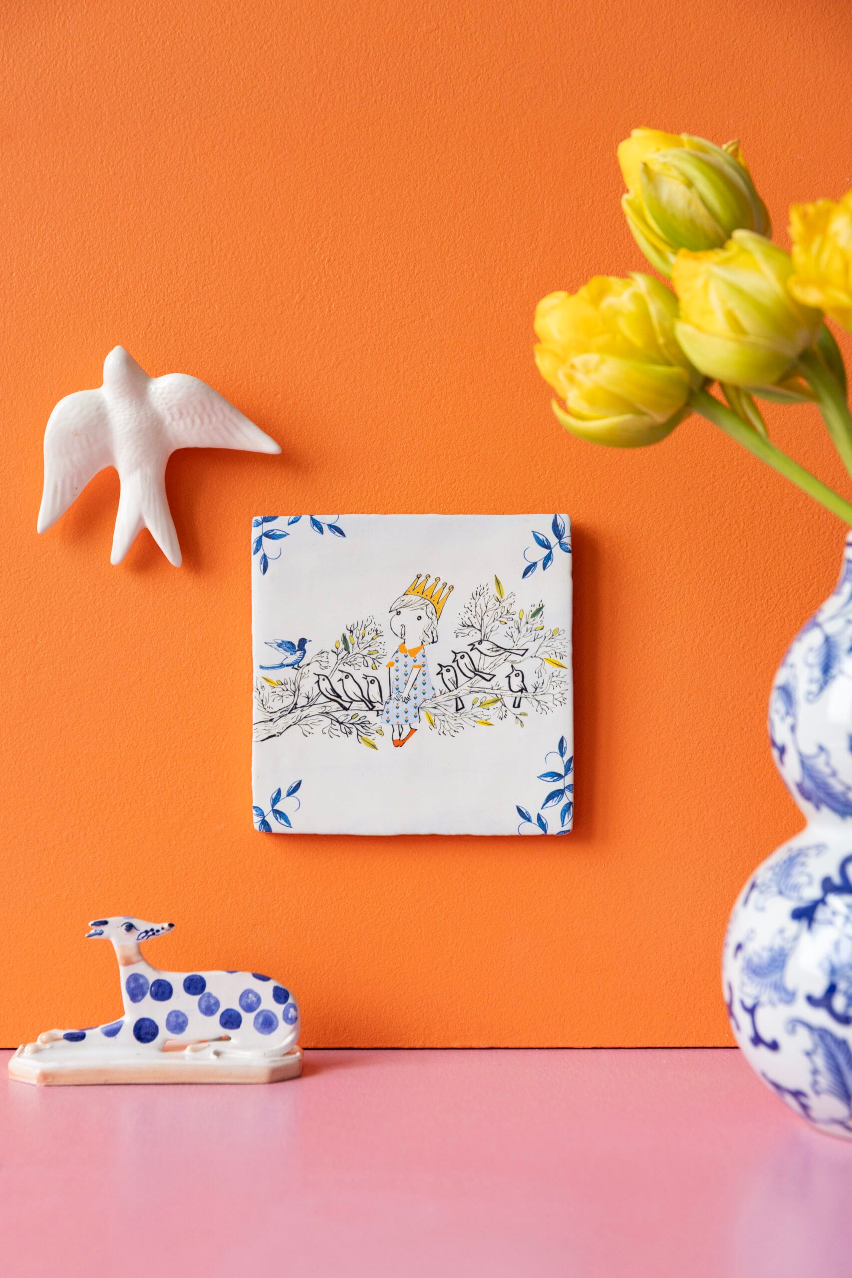 Tegel genaamd Vogelkoortje met illustratie van een gekroond meisje tussen vogels op een oranje muur, omringd door keramiek en gele tulpen, StoryTiles x Fiep Westendorp