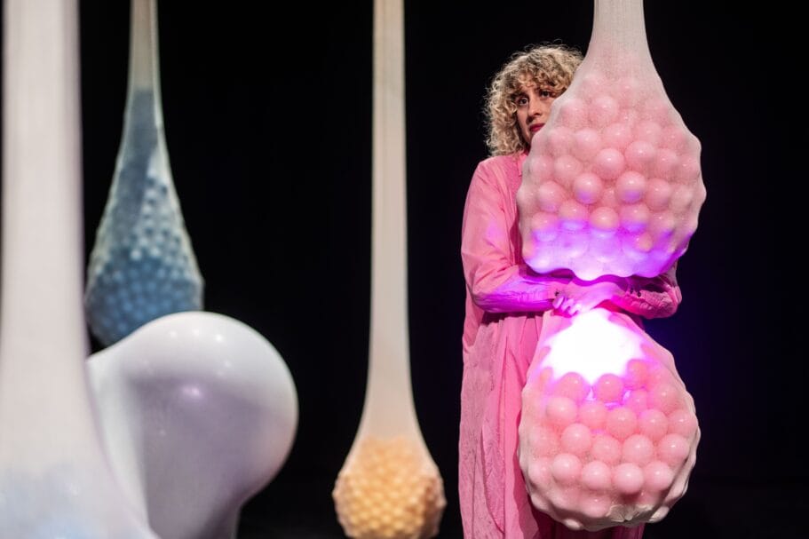 Theatermaker Karlijn Kistemaker in de voorstelling Buikzwam, over endometriose, in een roze gewaad omhelst de actrice een grote hangende zak vol roze oplichtende bollen op een donker podium