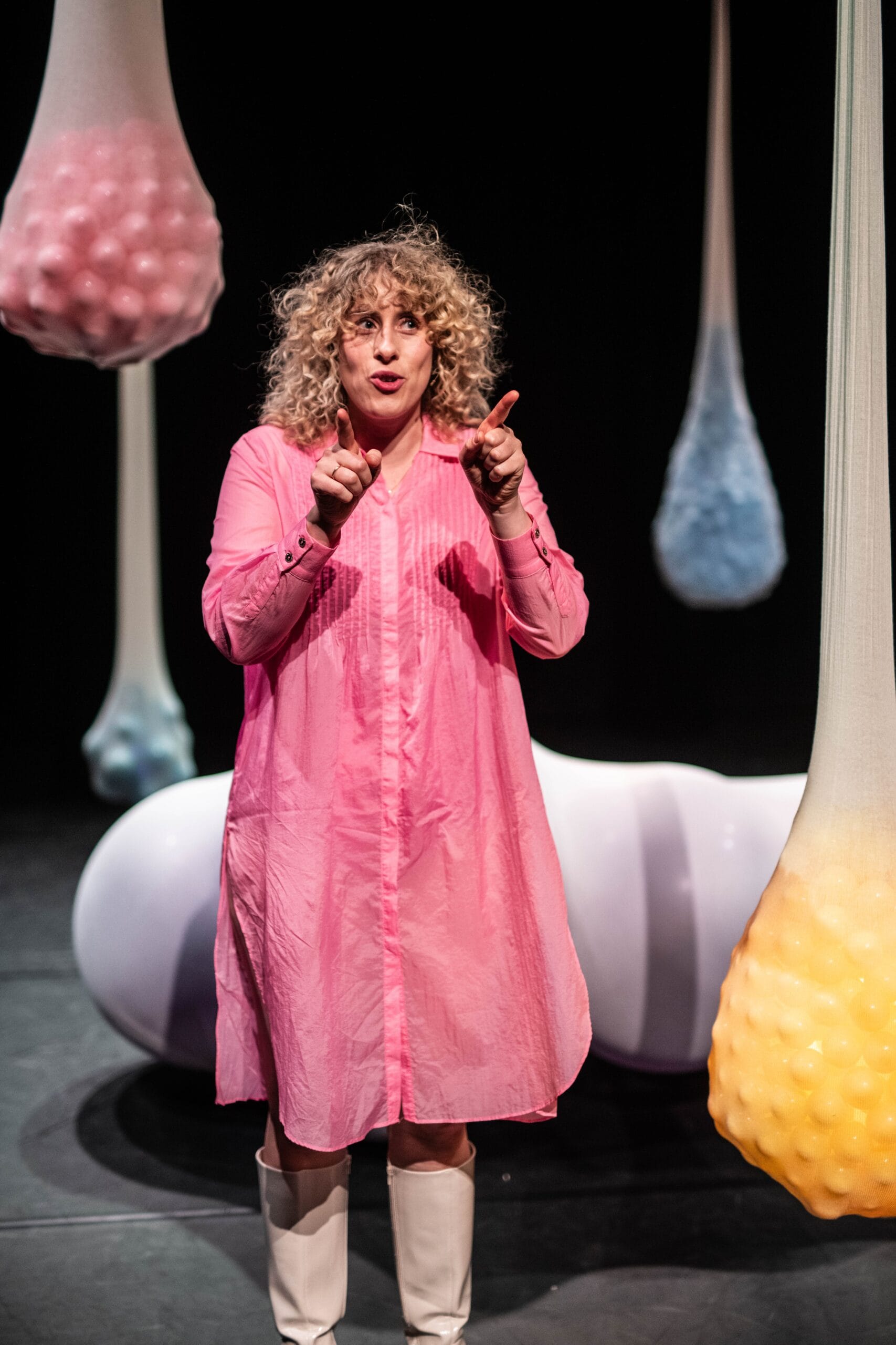 Theatermaker Karlijn Kistemaker in de voorstelling Buikzwam, over endometriose, vrouw met krullen in een roze jurk wijst omhoog op een podium tussen hangende, kleurrijke vormen